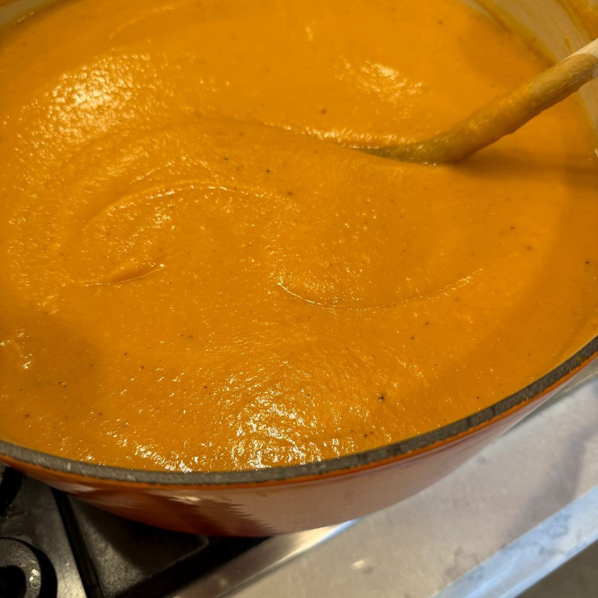 A Le Creuset pot of silky orange roasted butternut squash soup with a wooden spoon