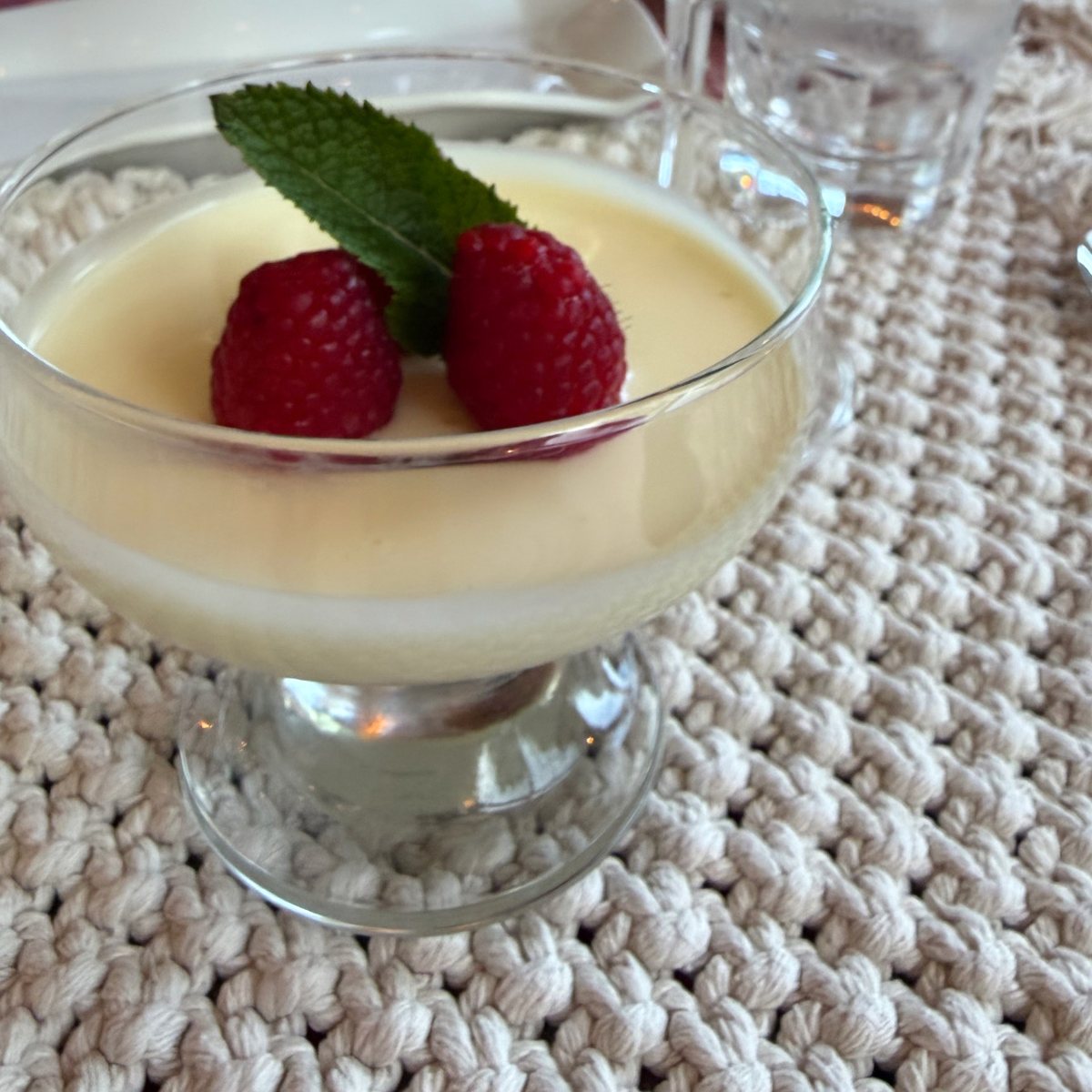 Lemon posset in a glass coupe topped with two raspberries and a mint leaf, a glass of white wine behind