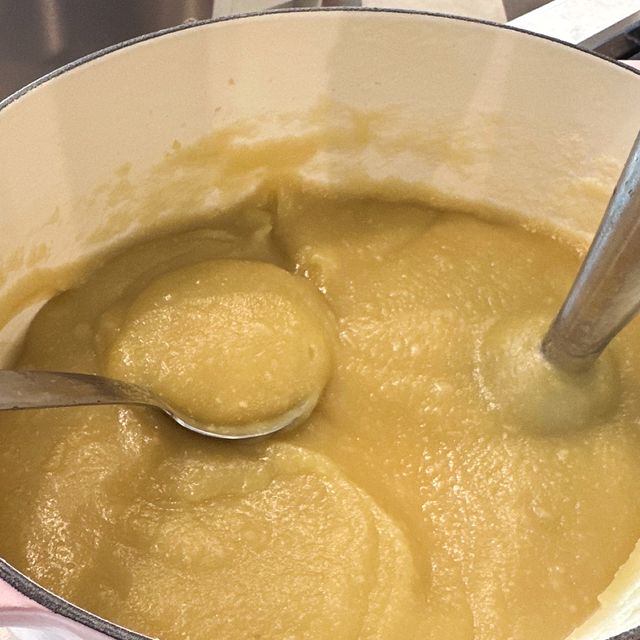 A pink Dutch oven of smooth homemade applesauce with a ladle and an immersion blender