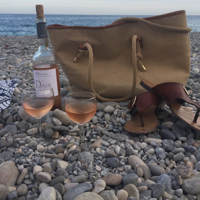 A bottle of Provence rosé, two glasses, a canvas tote and sandals on a pebble beach in Nice