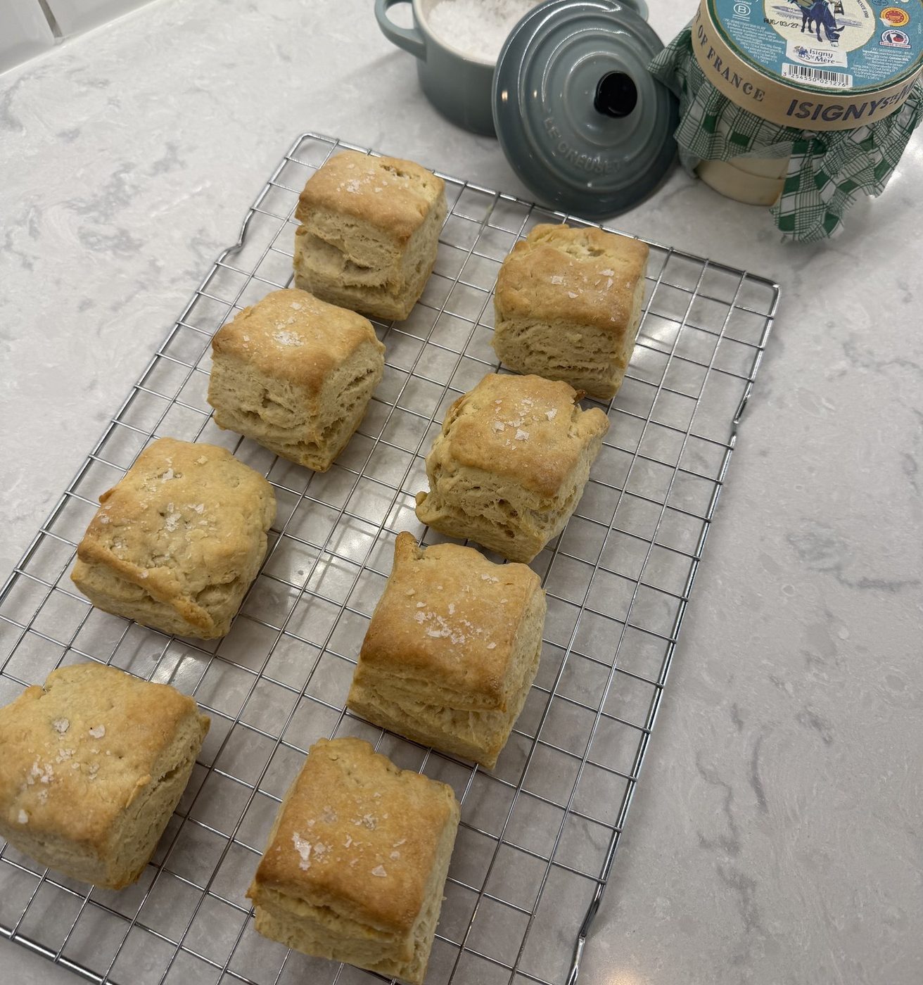 Square sour cream biscuits topped with flaky salt on a cooling rack, a tub of Isigny butter behind