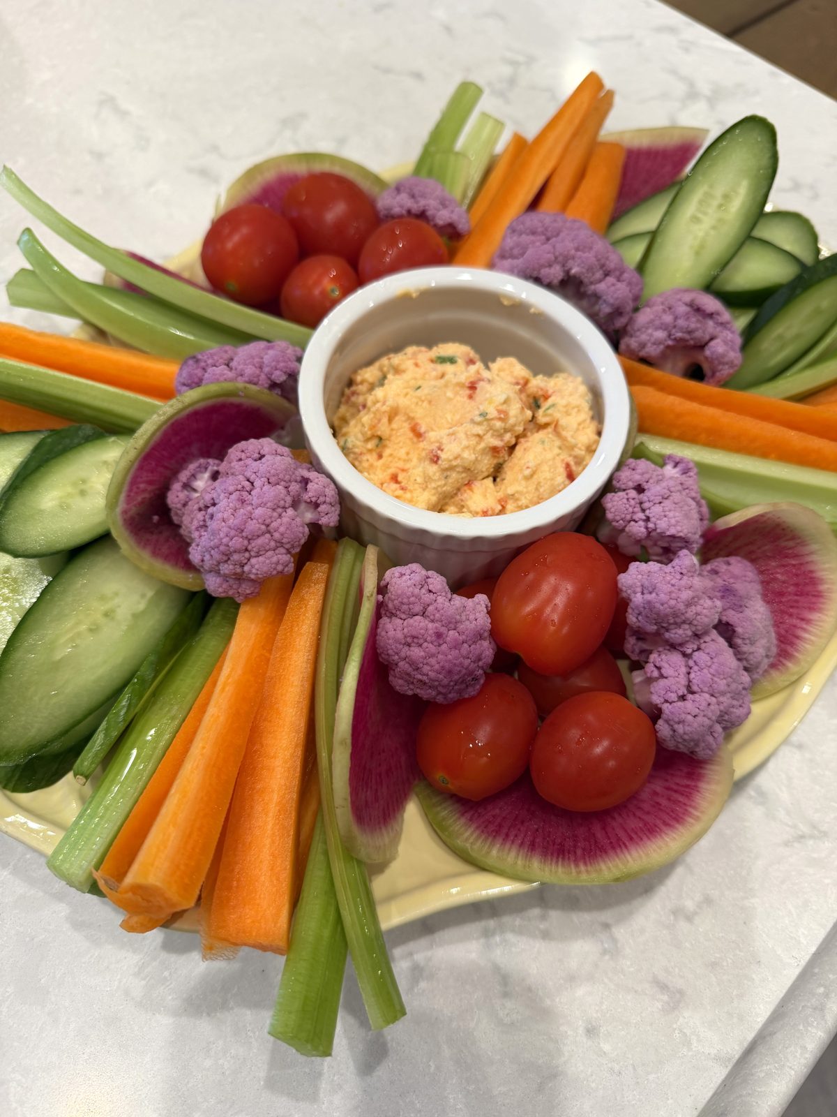 A ramekin of whipped pimento ricotta dip in the middle of a platter of carrots, celery, cucumber, cherry tomatoes, watermelon radish and purple cauliflower